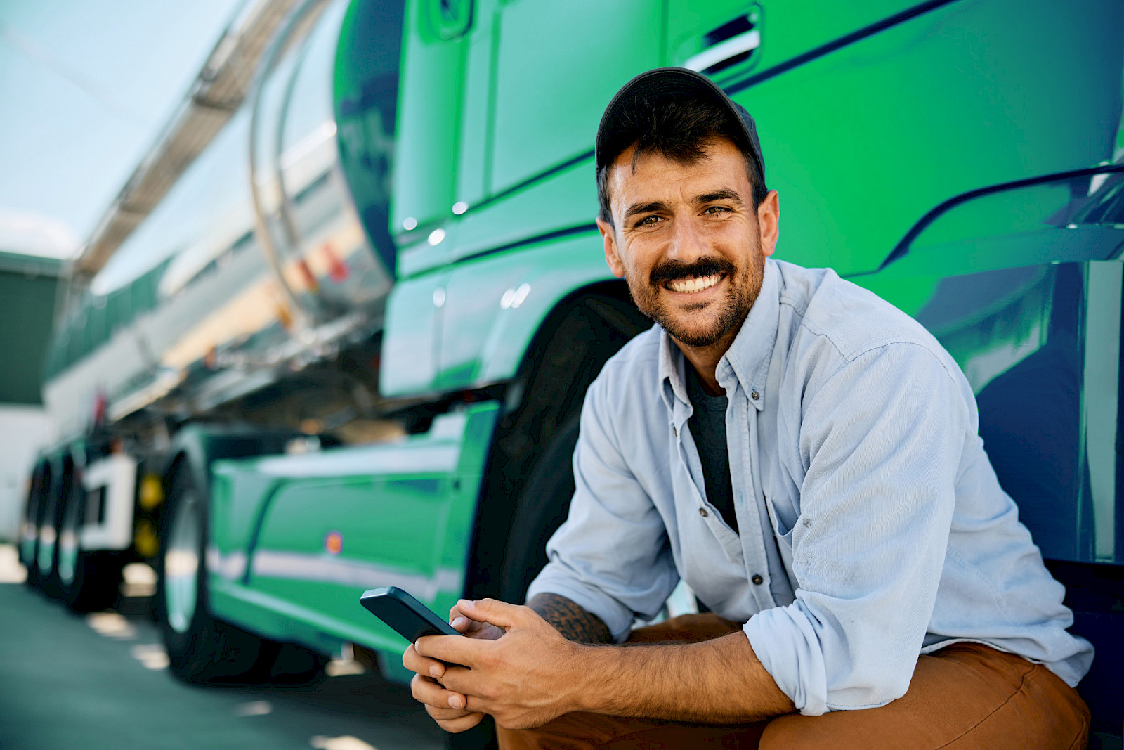 Lächelnder Lkw-Fahrer mit Smartphone vor einem grünen Tanklaster
